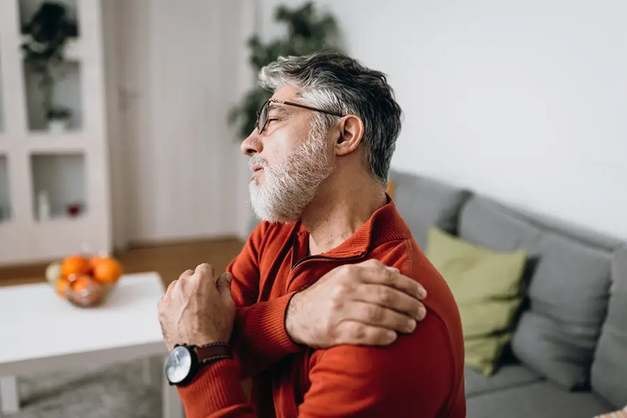 Older man with gray hair and beard sitting at home, holding his shoulder in discomfort, suggesting shoulder pain or stiffness.